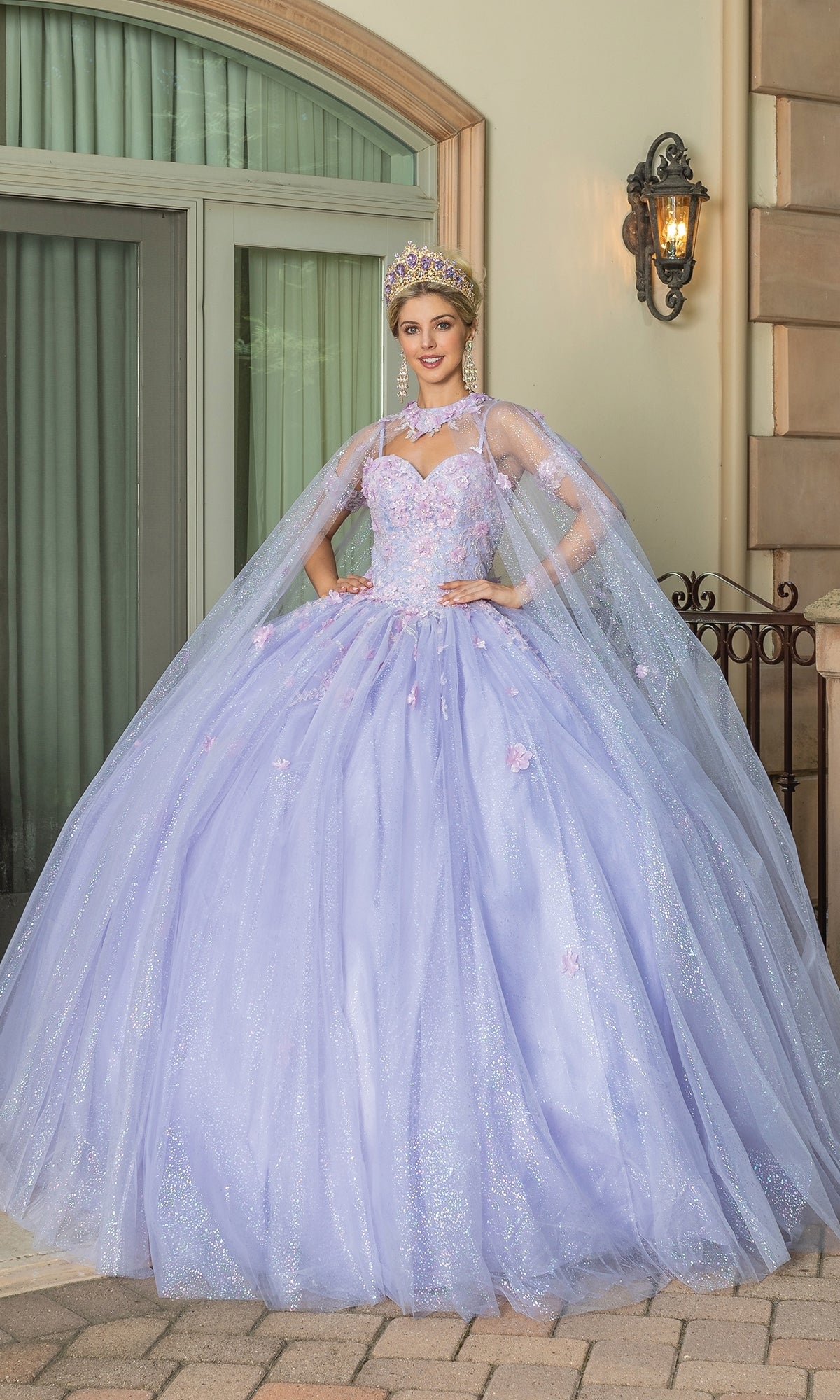 A woman stands outside wearing the Quinceanera Dress 1785 By Dancing Queen—a sparkling lilac ball gown with a full skirt, matching cape, jeweled tiara, and hands on hips.